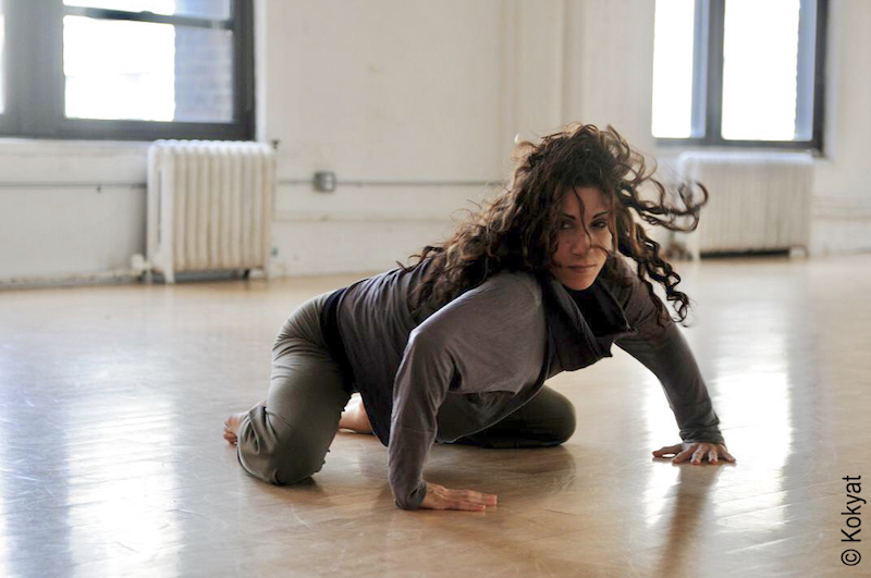 A woman crouches on the floor in grey dance clothes in a studio.